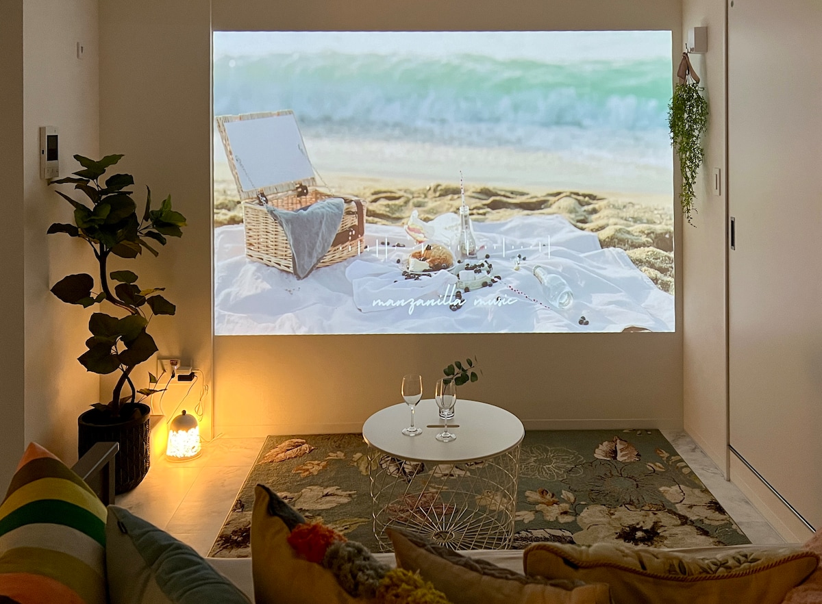 A cozy living area features a large 122-inch projector displaying a serene beach scene. A low round table with two wine glasses sits on a textured rug, with colorful cushions adorning a comfortable sofa. Soft lighting contributes to the inviting ambiance.