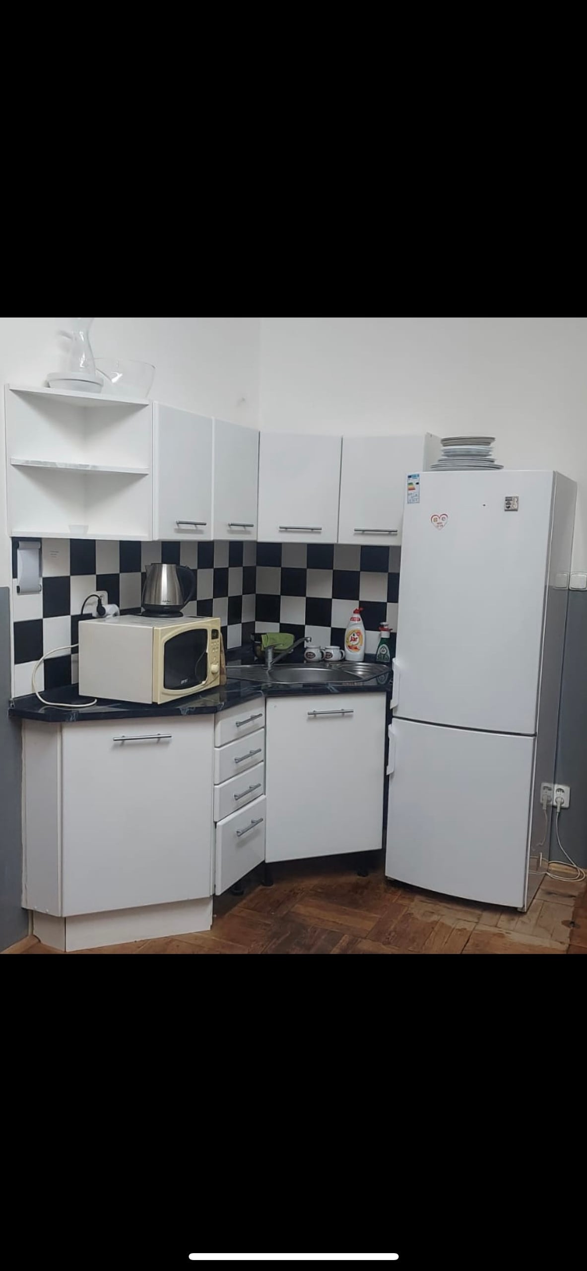 A compact kitchen features a mix of white cabinetry and checkered black and white backsplash. Essential appliances include a microwave, refrigerator, and kettle, with countertop space utilized for food preparation. Shelves above hold dishware and other kitchen items, enhancing functionality.