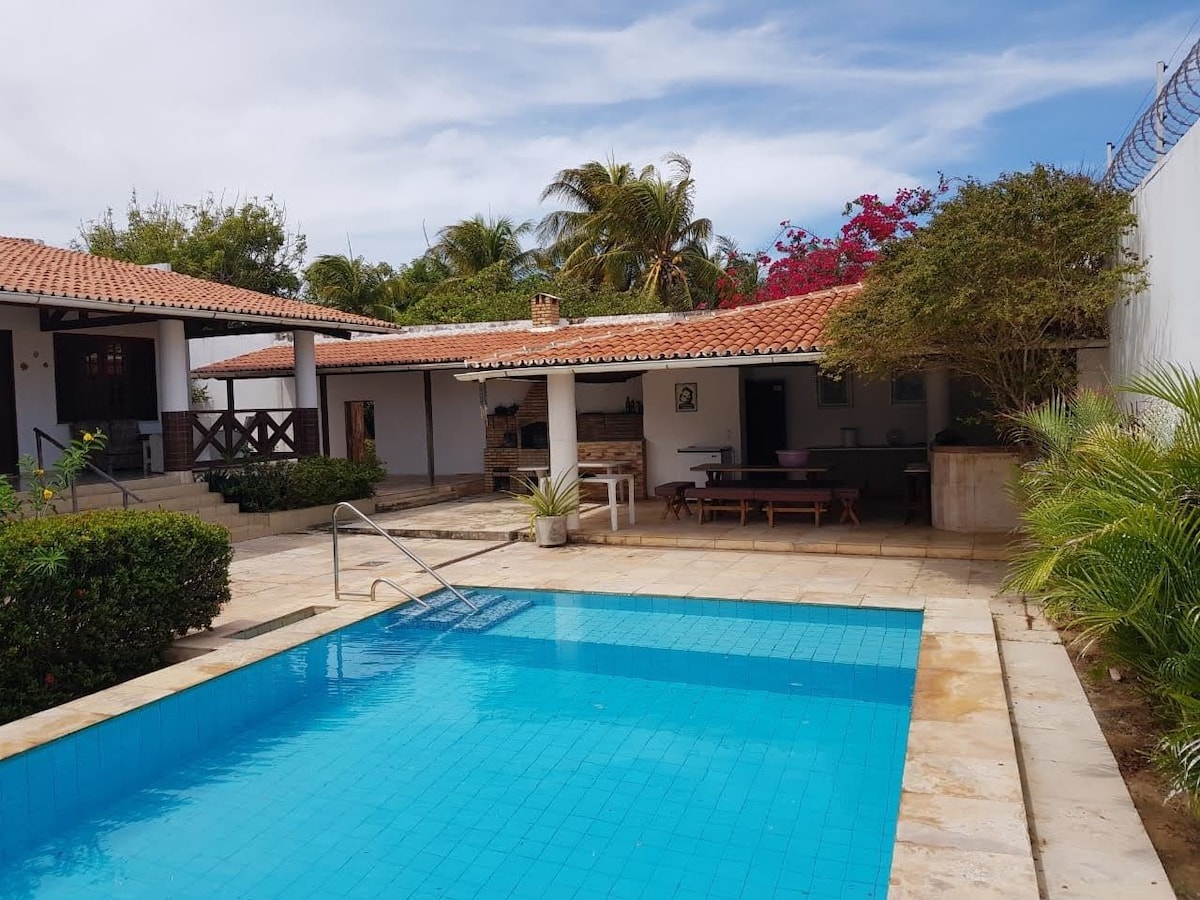 A refreshing swimming pool is set in a stone-tiled area surrounded by lush greenery and flowering plants. The pool area provides easy access to a covered outdoor dining space featuring a long table and chairs. The structure's roof is adorned with terracotta tiles.