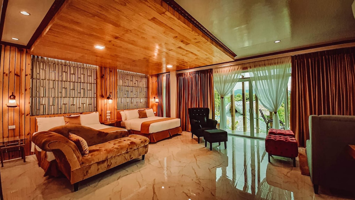 An inviting room features two spacious beds with neutral bedding, complemented by a dark armchair and a small ottoman. Floor-to-ceiling windows allow natural light to fill the space, while the wooden ceiling and accent lighting create a warm ambiance. Elegant curtains frame the view outside.