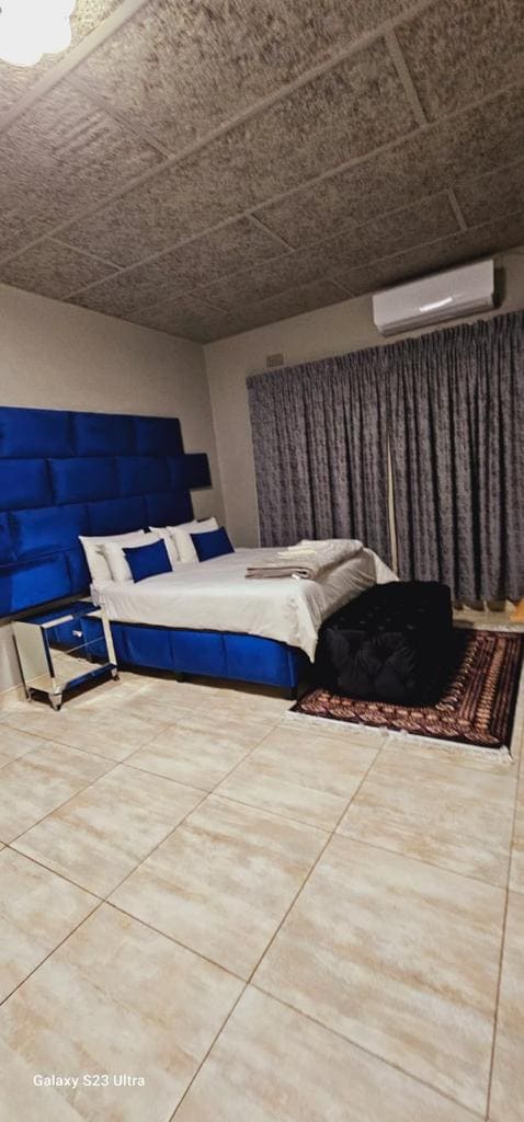 A spacious sleeping area features a large blue upholstered headboard and light-colored bedding. Soft lighting illuminates the room, which includes a neatly arranged bedside table and a dark seating area with a plush ottoman. Floor-to-ceiling curtains frame the window, adding to the cozy ambiance.