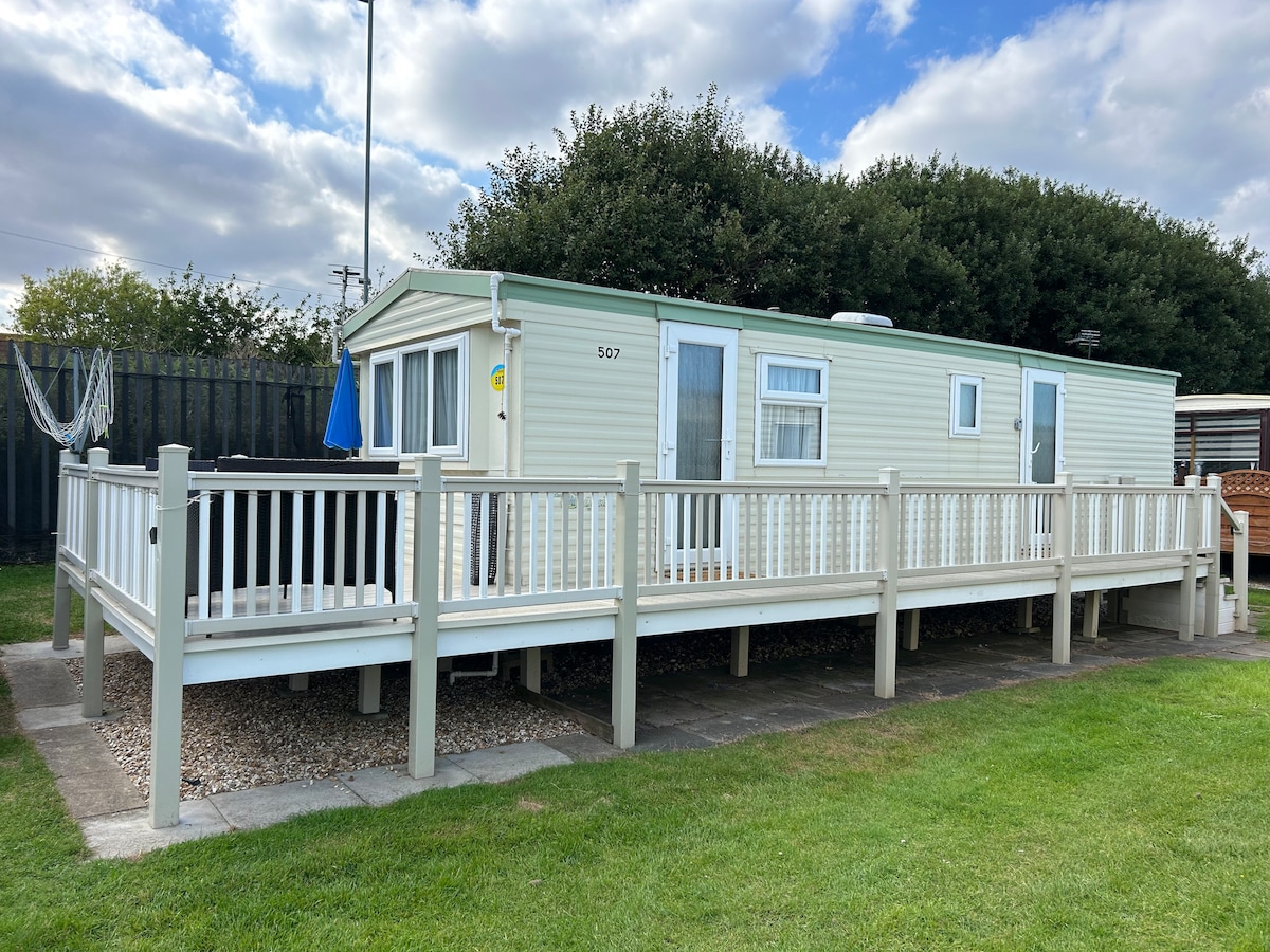 A light-colored caravan is situated on a grassy area with a spacious outdoor gated veranda. The exterior features multiple windows, providing ample natural light. Behind the caravan, a shaded area with trees is visible, contributing to a serene environment.