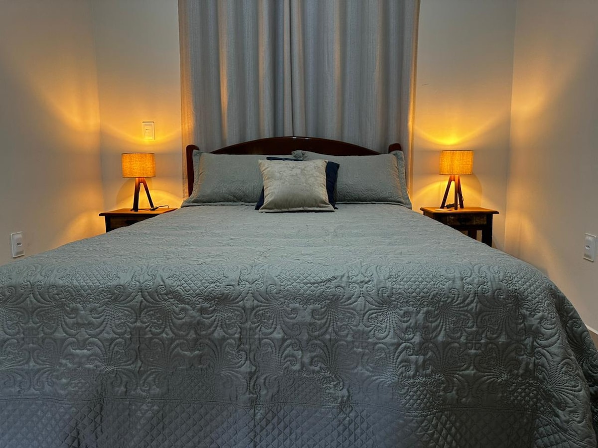 A comfortable sleeping area is presented with a spacious bed adorned with a textured quilt. Warm light is emitted from two bedside lamps, creating a cozy ambiance. Neutral-colored curtains frame a window, complementing the soothing tones of the room.