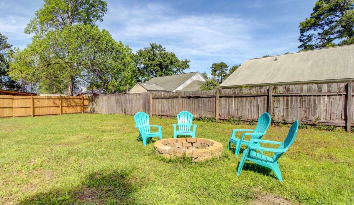 A spacious backyard is revealed, featuring a circular fire pit surrounded by four blue Adirondack chairs. Lush green grass covers the area, complemented by a tall tree and wooden fencing providing privacy. Nearby structures are partially visible in the background.