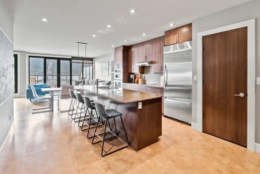 A modern kitchen features sleek wooden cabinetry and stainless steel appliances. An island with seating for five provides ample space for meal prep. Natural light floods the area through large windows, highlighting a dining table with chairs, adjacent to the kitchen.