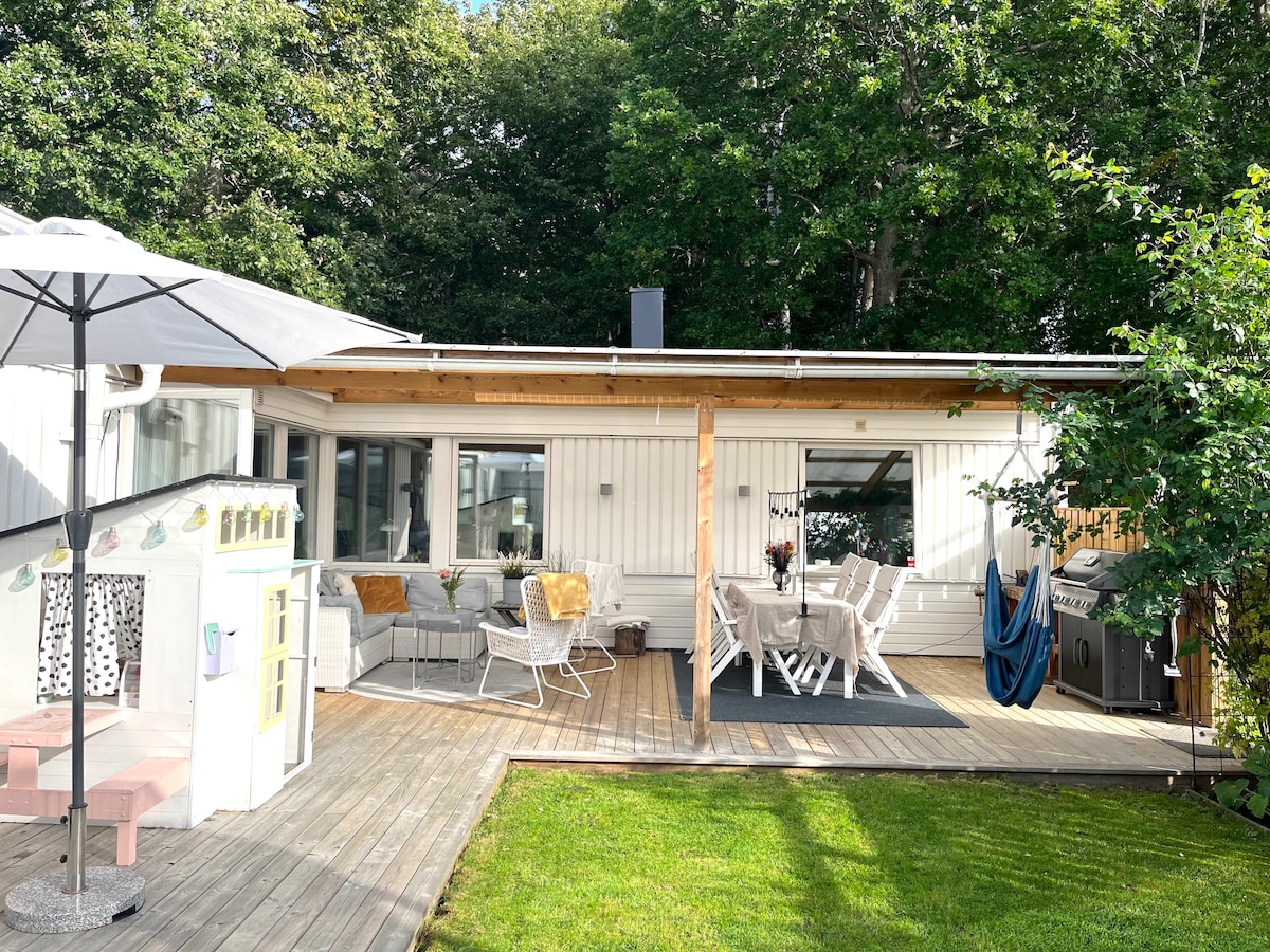 The image displays a bright, single-story house featuring a spacious wooden deck with outdoor seating and a picnic table. A gas grill is positioned nearby, and a playhouse can be seen in the garden, surrounded by lush greenery.