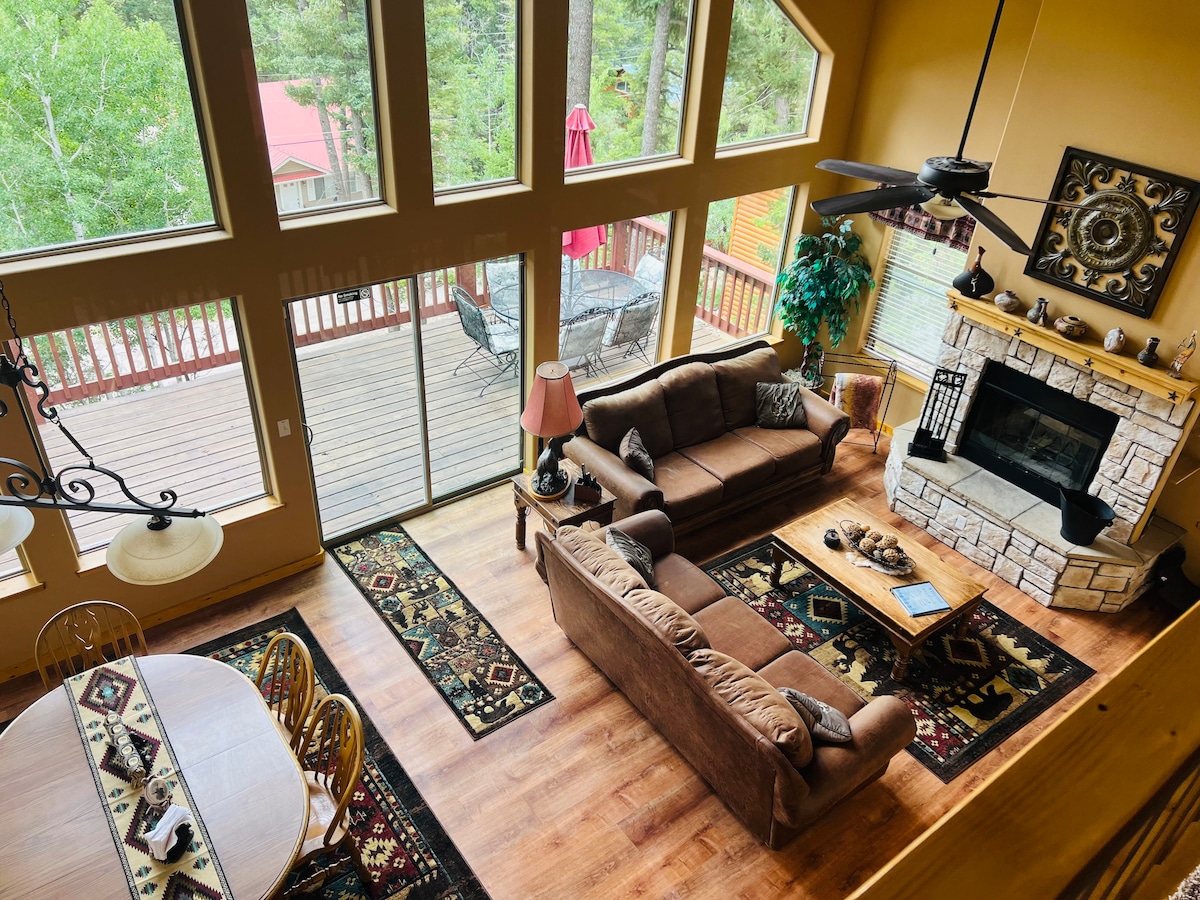 An open living area showcases a large, comfortable sofa facing a stone fireplace. Abundant natural light enters through tall, framed windows, illuminating the wood floors and area rugs. A dining table with surrounding chairs is visible, while a deck area with a table and umbrella extends outdoors.