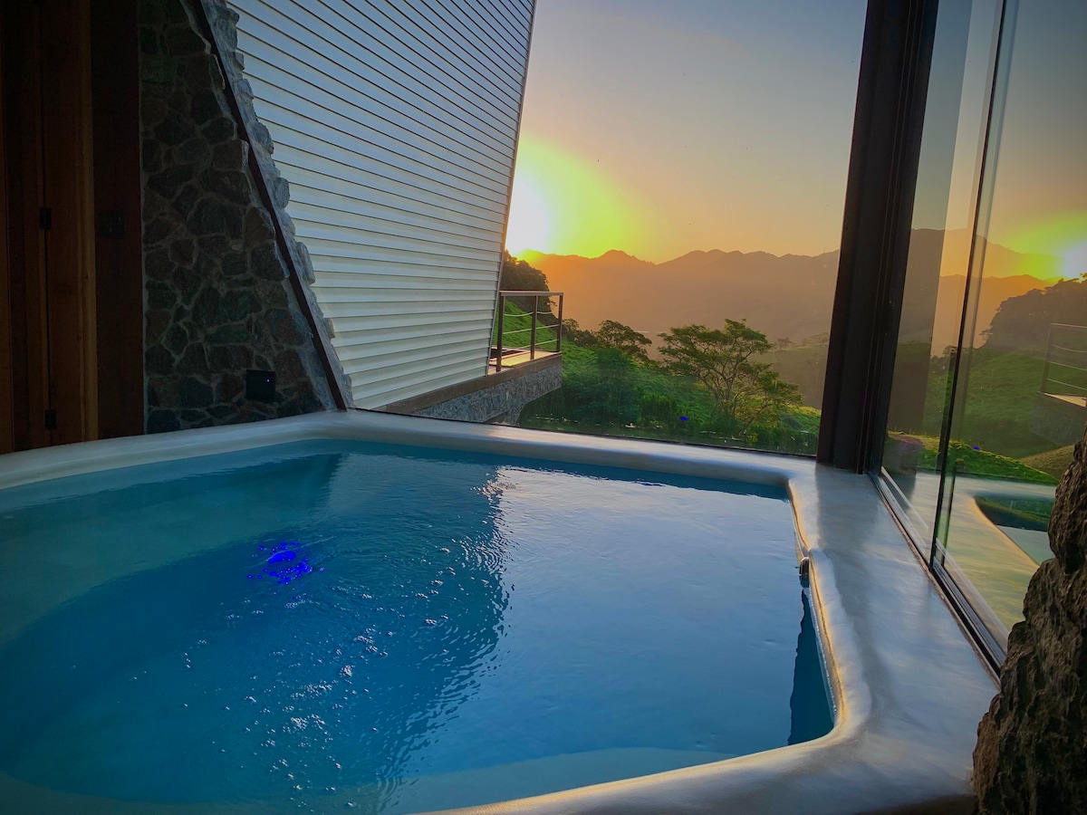 A Mediterranean-inspired mini-pool is highlighted, showcasing clear water and ambient lighting. The view reveals a stunning sunset over the mountains, with large glass windows offering a seamless connection to nature.