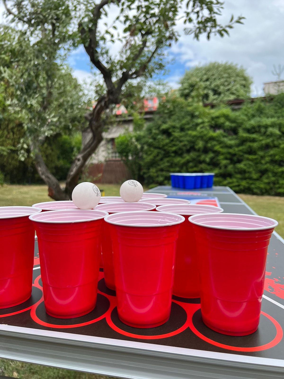 A gaming setup is presented outdoors with red plastic cups arranged in a triangle formation. Two white ping pong balls are positioned nearby. A blue cup is situated further back on the table, surrounded by lush greenery and a few trees in the background.