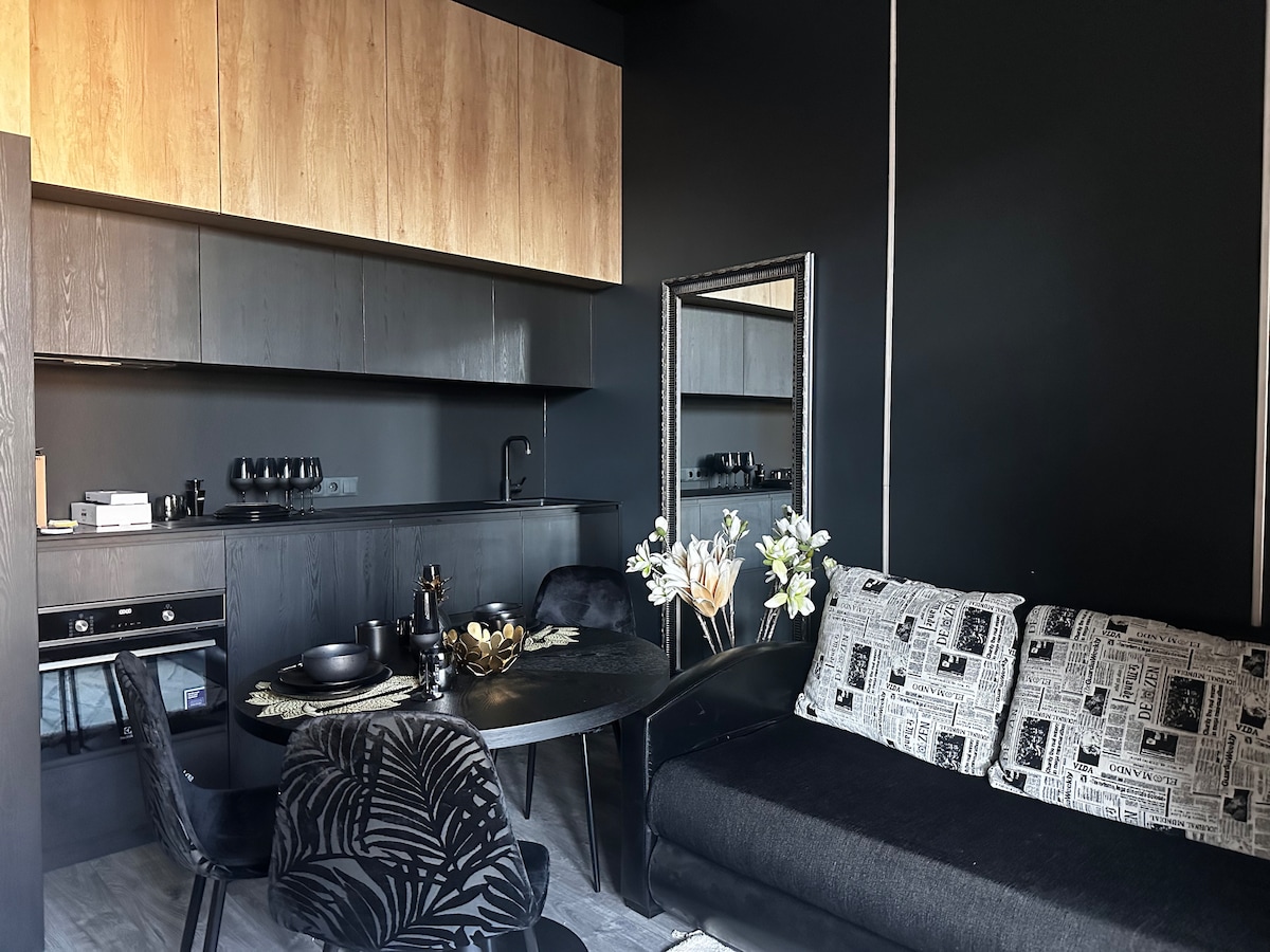A modern kitchen area is seen, featuring sleek wooden cabinetry and dark countertops. A round dining table with stylish chairs is set, while decorative flowers add a touch of elegance. A cozy seating area with a patterned sofa provides comfort, with a mirror enhancing the space.