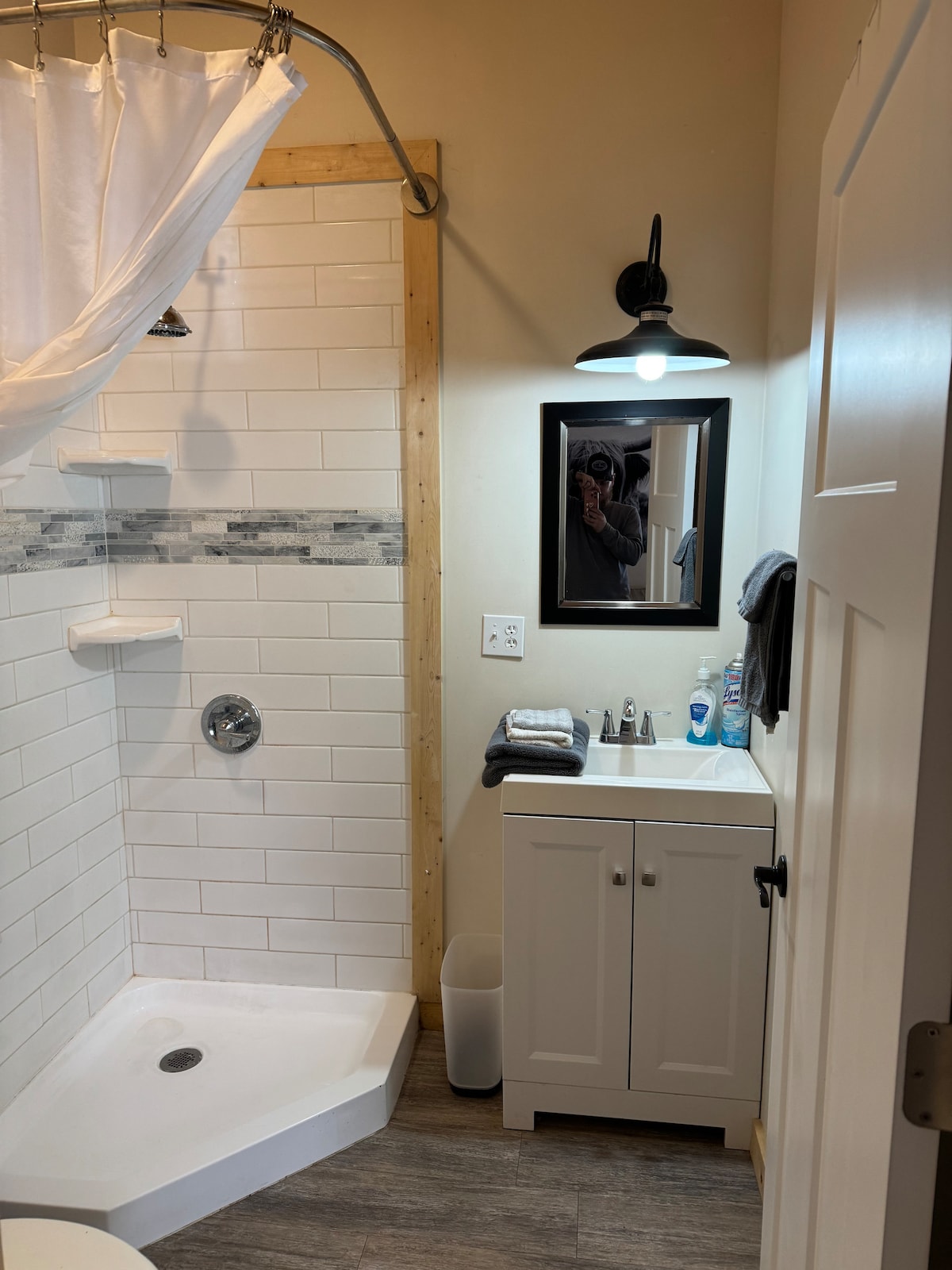 A clean bathroom is presented, featuring a corner shower with a glass door and a modern fixture. A small white cabinet holds neatly stacked towels and toiletries. A mirror above the sink reflects soft lighting, and the tile walls are accented with a decorative strip.