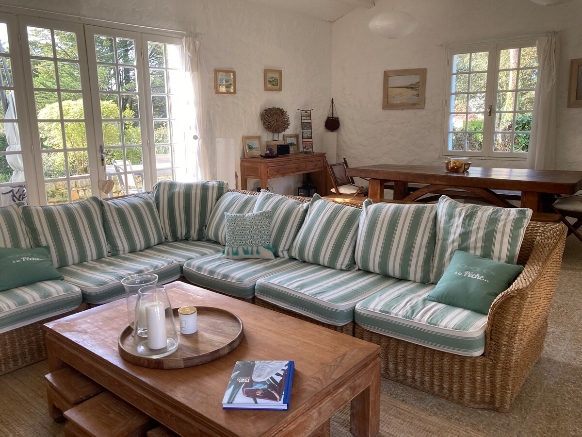 A spacious living area is highlighted by a large, L-shaped sectional sofa featuring striped cushions. A wooden coffee table sits at the center, complemented by a decorative tray and a soft pitcher. Sunlight filters through multiple windows, creating a bright and airy environment.