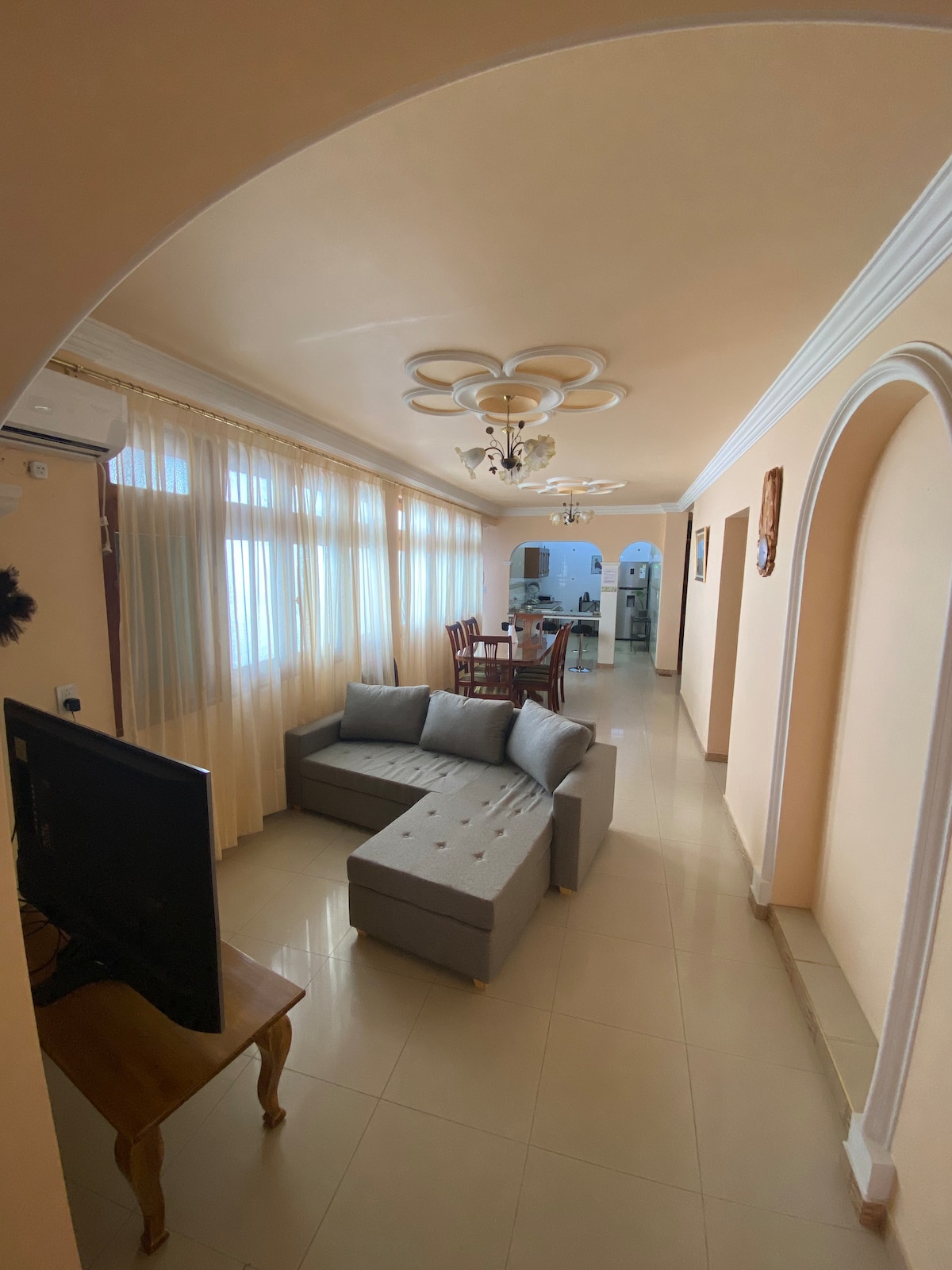 A bright living area features a modern sectional sofa and a wooden table with chairs. Large windows are adorned with sheer curtains, allowing natural light to fill the space. An arched entryway leads towards the dining area and a glimpse of the kitchen.