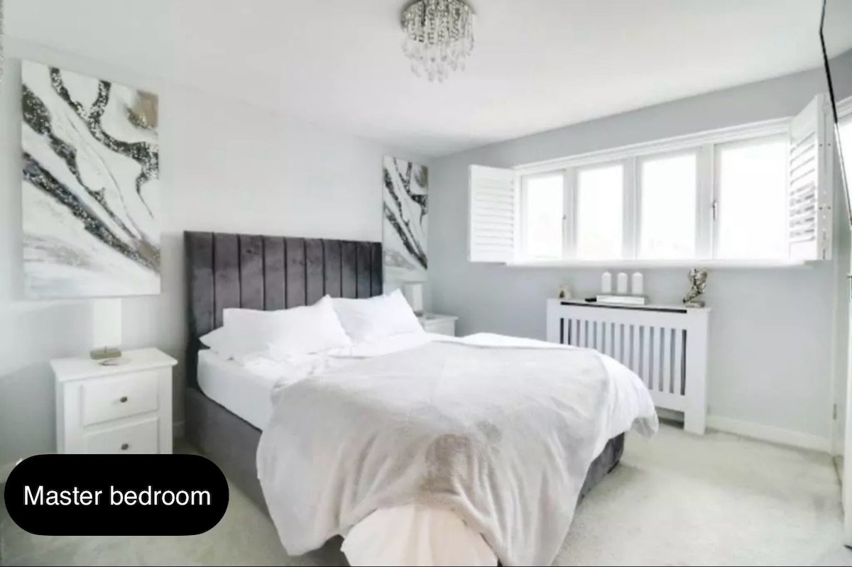 The master bedroom features a spacious bed dressed in soft linens. Two light-colored artworks adorn the walls, and a stylish chandelier hangs from the ceiling. Shutters allow for adjustable natural light, while a small radiator sits beneath the window.