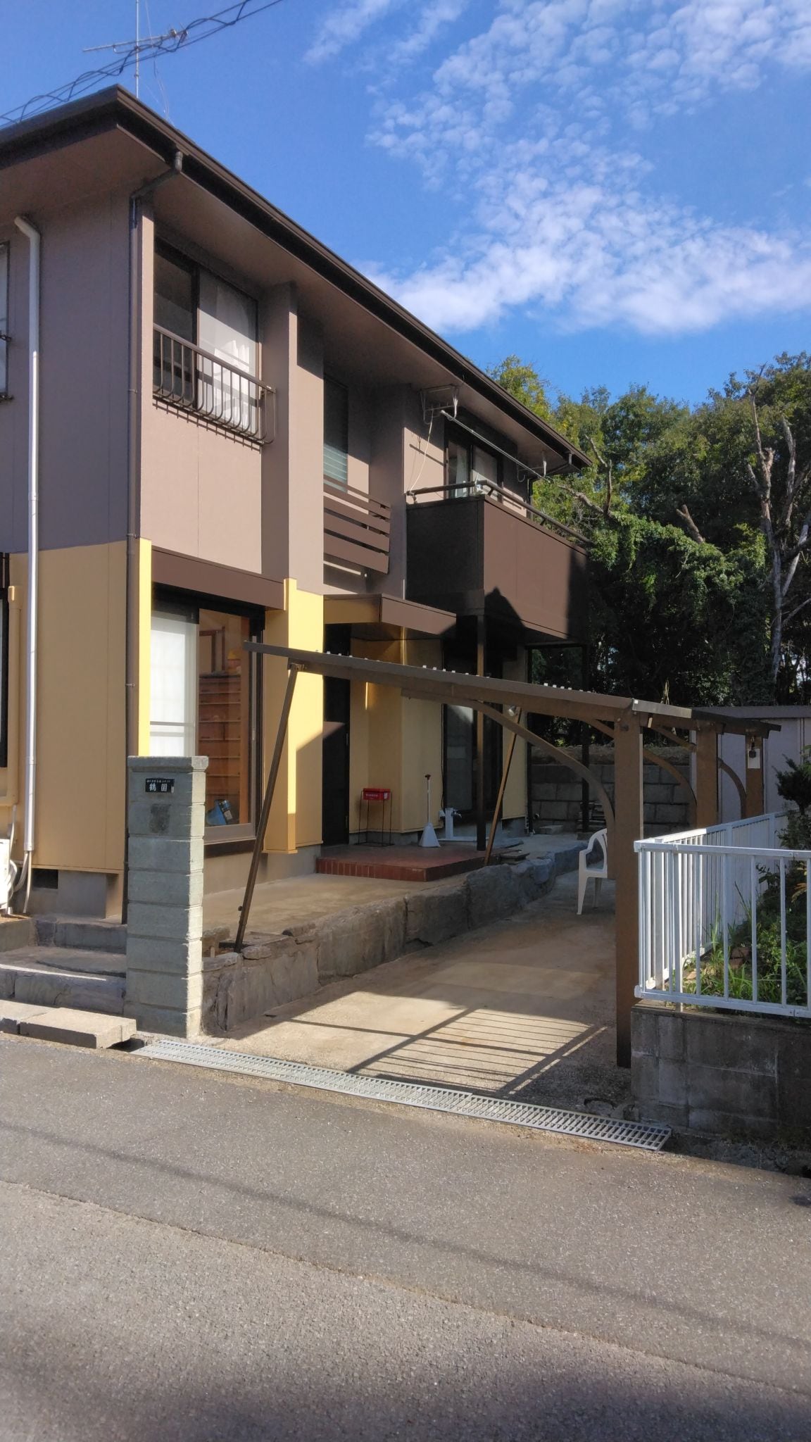 A two-story house is presented, featuring a mixture of brown and yellow exterior walls. A concrete walkway leads to the entrance, which is framed by a wooden awning. Lush greenery is visible in the background, enhancing the home's inviting appearance.