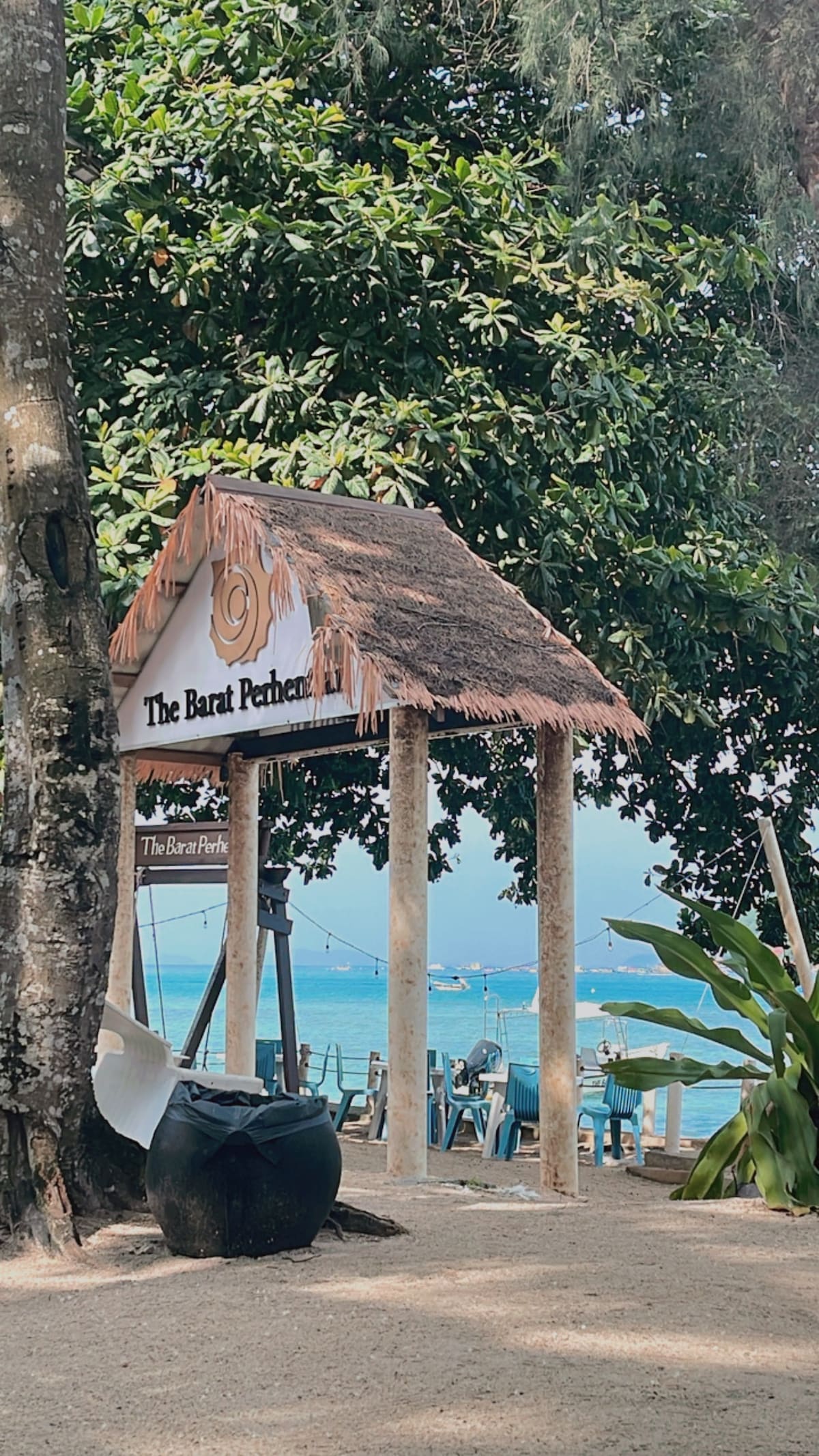 A thatched-roof structure marks the entrance to The Barat Perhentian, surrounded by lush greenery. The sandy path leads toward a serene beach view, with distant boats visible on the calm water. The setting invites relaxation in a coastal environment.