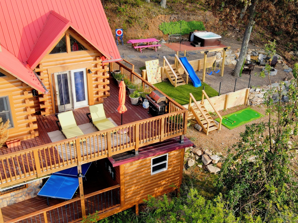An aerial view of the cabin's outdoor area reveals a spacious deck with lounge chairs and an umbrella. A children's playset, a hot tub, and a putting green are visible in the yard, surrounded by trees, highlighting a perfect retreat for relaxation and recreation.