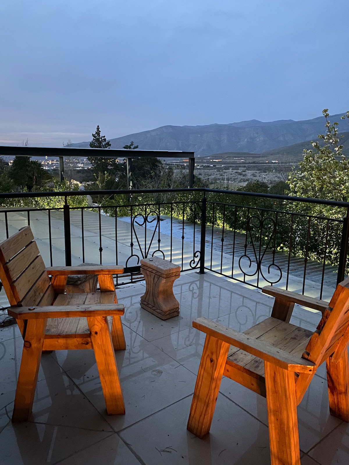A balcony area features two wooden chairs and a small table, positioned to enjoy views of distant mountains and the surrounding landscape. The evening sky casts a soft light, highlighting the inviting outdoor seating space.