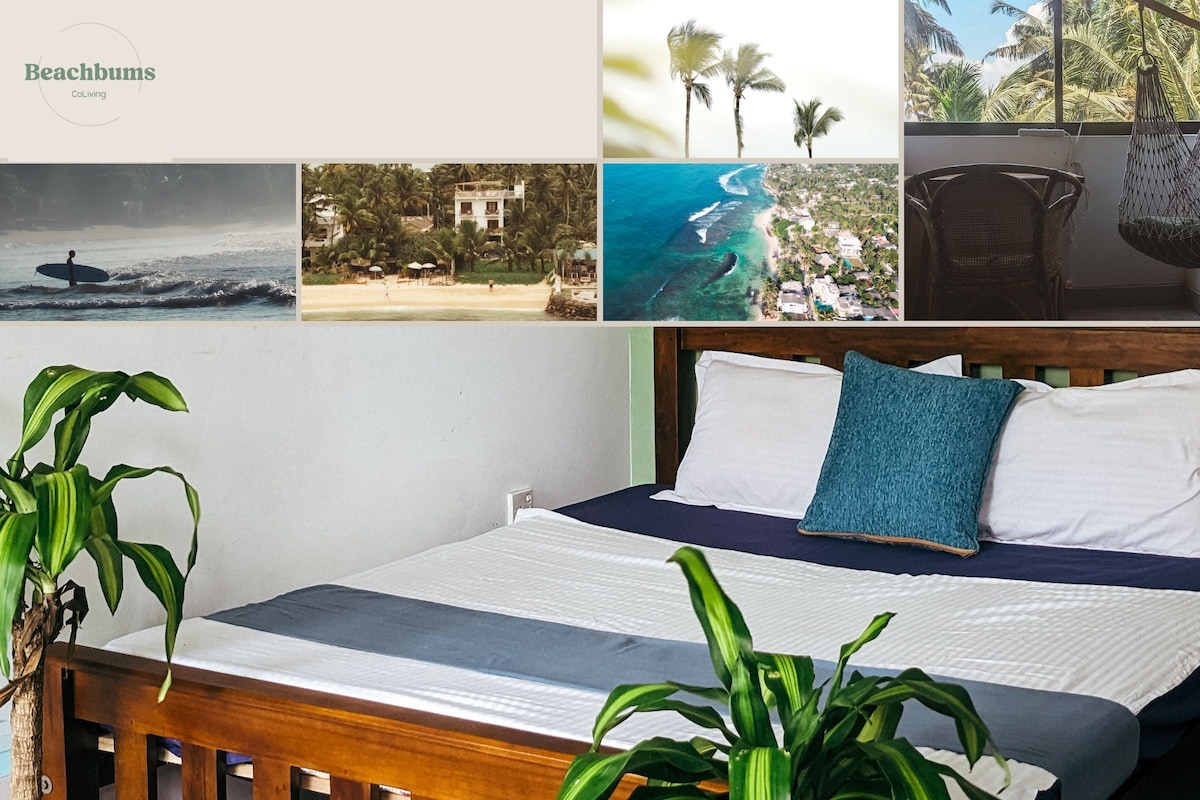 A spacious bed is centered in the room, dressed in crisp white linens with a textured blue accent pillow. Lush green plants are positioned by the bedside, contributing to a relaxed ambiance. Light filters in, highlighting the natural materials and tranquil color palette.