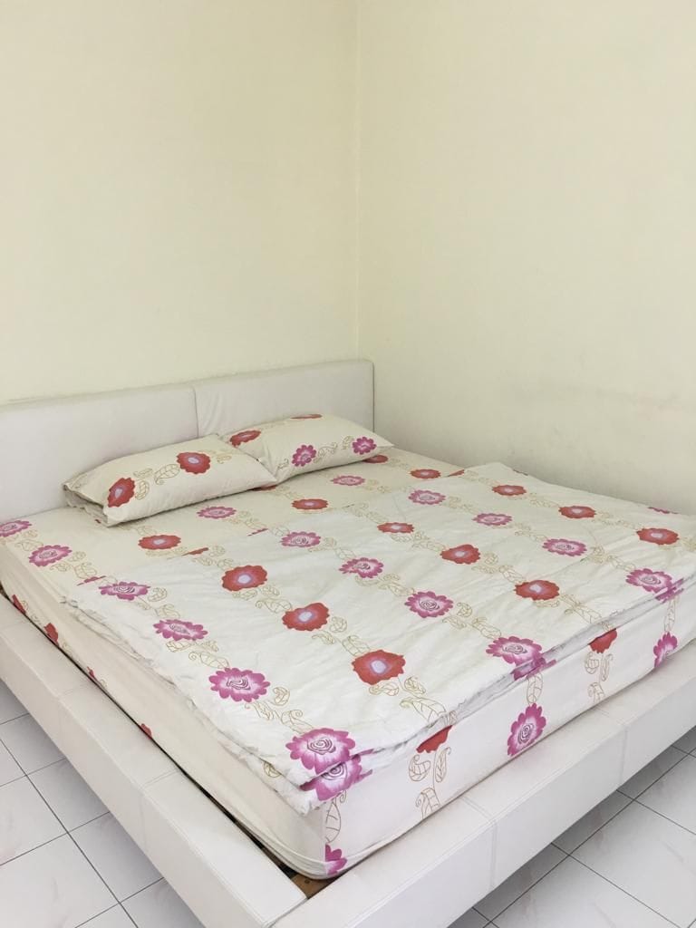 A spacious bed is presented, covered with a floral-patterned comforter featuring pink and white hues. Two pillows rest at the head of the bed. The bedroom walls are minimally styled, providing a neutral backdrop to the bedding.