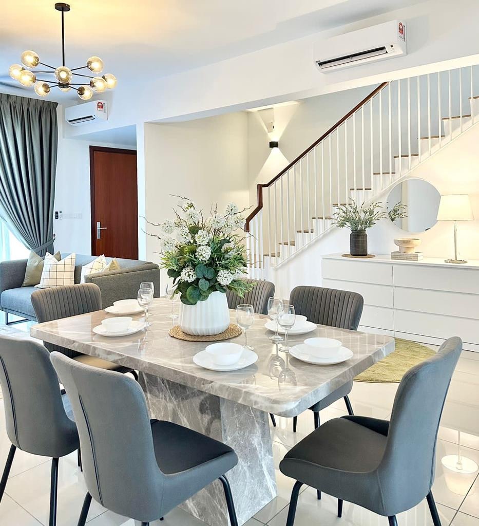 A spacious dining area features a marble table set for a meal with plates and glasses. Elegant chairs surround the table. In the background, a cozy living room is visible, adorned with decorative cushions and modern lighting fixtures.