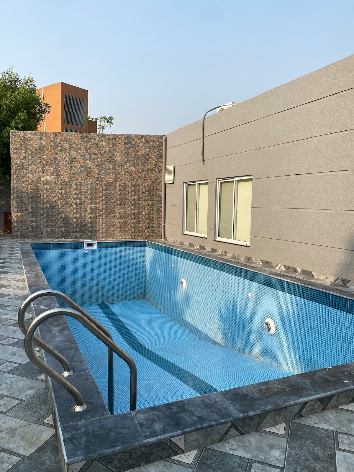 A private swimming pool is featured with a clear blue mosaic tile surface and built-in steps for easy access. The surroundings include a textured wall and several windows providing natural light, while decorative tiles create a patterned border around the pool area.