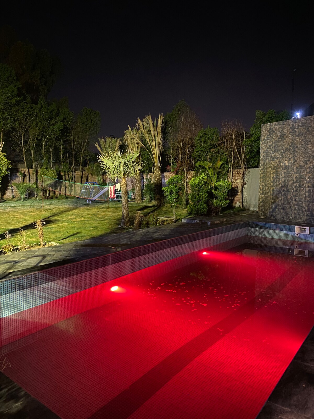 A private swimming pool is illuminated with soft red lighting, creating a serene atmosphere at night. Surrounding greenery includes various plants and trees, contributing to a tranquil outdoor space that enhances relaxation.