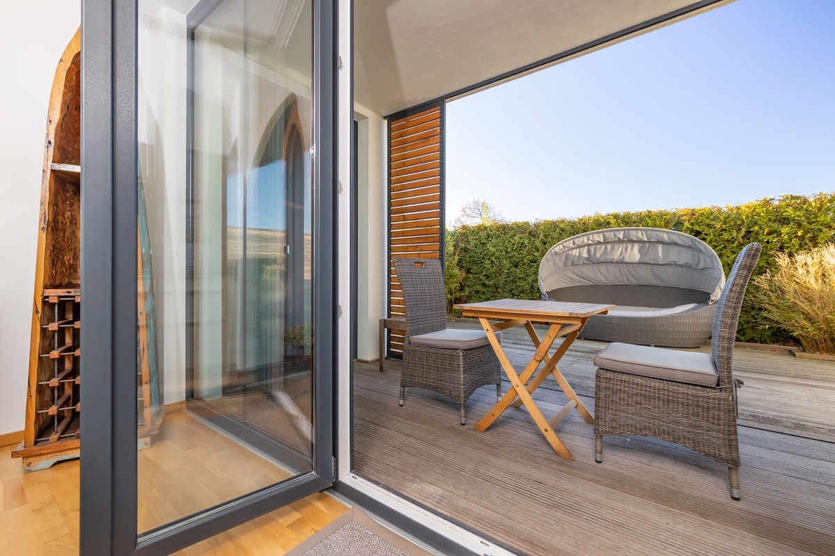 An outdoor terrace features a cozy Muschelloungebett and a wooden table surrounded by wicker chairs. The space is framed by low greenery and is bathed in natural light, offering a serene setting for relaxation.