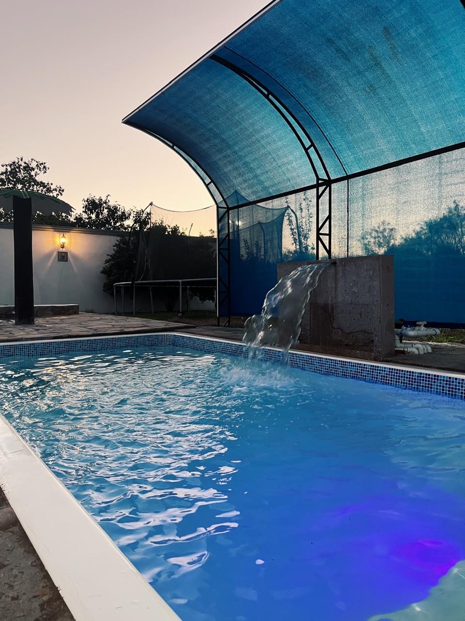 A swimming pool is visible, featuring clear blue water with soft ripples. Above the pool, a large, blue canopy provides shade. A gentle waterfall flows from a stone feature into the pool, enhancing the serene environment.