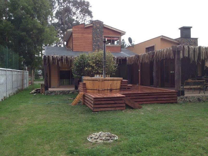 A wooden tub is positioned on a raised deck within a spacious backyard, surrounded by greenery. The house features a unique blend of natural materials, with a combination of stone and wooden elements visible. The patio area is designed for relaxation and outdoor dining.