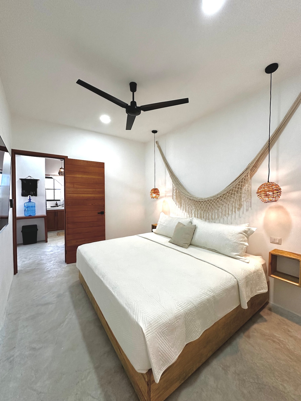 A bright bedroom features a spacious bed with a white blanket, complemented by decorative hanging elements. A ceiling fan enhances airflow, and wall sconces provide ambient lighting. A doorway leads to an ensuite bathroom, visible in the background.