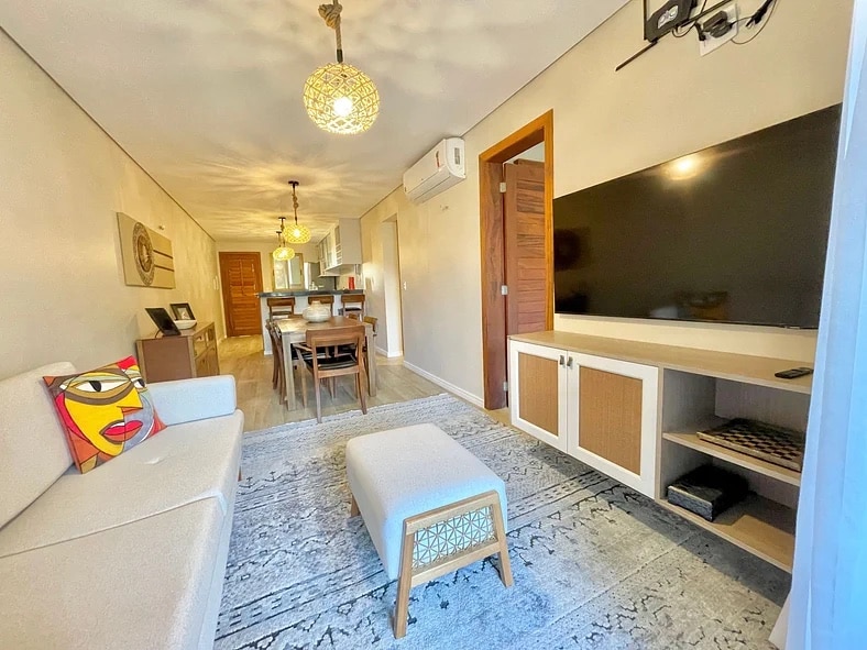 A well-lit living area features a neutral-toned sofa and an ottoman, complemented by a decorative cushion. A wooden dining table is visible in the background, along with a modern television mounted on a console. Warm pendant lights enhance the comfortable ambiance.