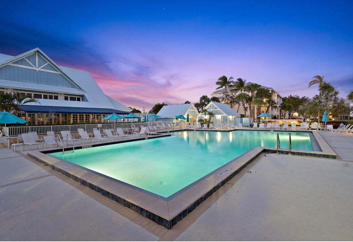 A well-maintained swimming pool is surrounded by lounge chairs and umbrellas, set against a colorful sunset sky. The pool area is spacious, showcasing calm, inviting waters that reflect the evening light, inviting guests to relax and enjoy the tranquil outdoor setting.