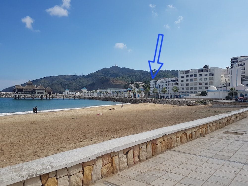 A scenic view of a sandy beach is framed by gentle waves lapping at the shore. In the background, a small hill rises, and several buildings are visible along the coastline, creating a harmonious blend with the landscape.