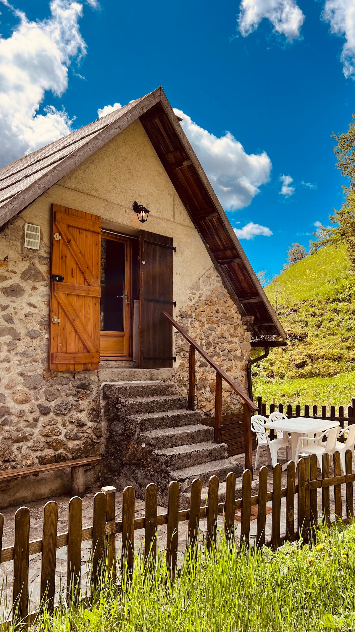 The exterior of the charming stone cottage is showcased with wooden shutters opened wide. A set of steps leads to the front door, framed by a wooden railing. A small patio with white chairs is visible, surrounded by a grassy area and a wooden fence.