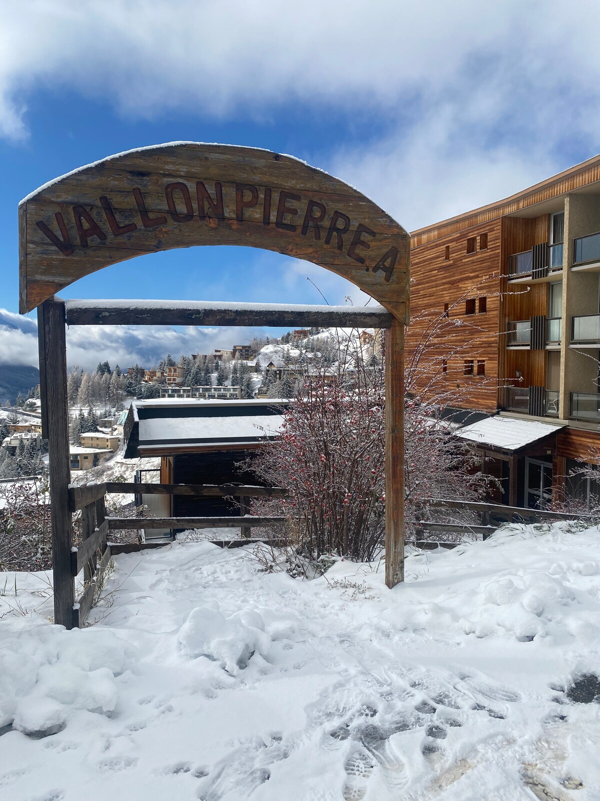 A wooden archway labeled 'Vallonpierre' stands at the entrance, leading into a snowy landscape. The scene features white snow covering the ground, with a multi-story building in the background, blending with the winter scenery and a few trees dusted in snow.