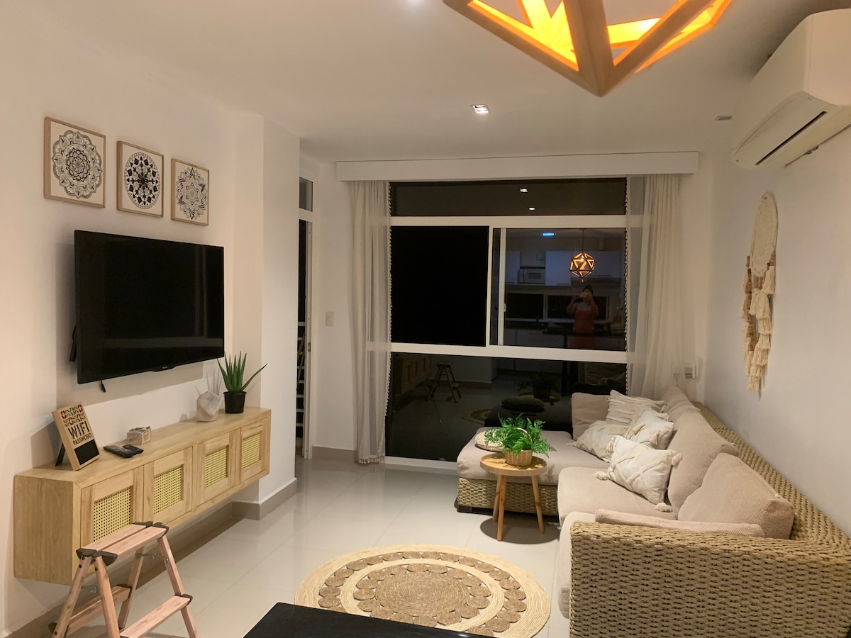 A cozy living area is featured with a light-colored sofa and a stylish coffee table. A television is mounted on the wall, while decorative elements include wall art and a woven wall hanging. Large windows provide natural light and a view of the surroundings.