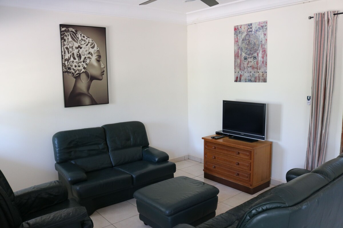 A living area is furnished with dark leather seating, including two armchairs and a sofa. A wooden TV stand supports a flat-screen television. A colorful wall art piece is displayed above the TV, while light-colored curtains on the windows allow natural light to enter the room.