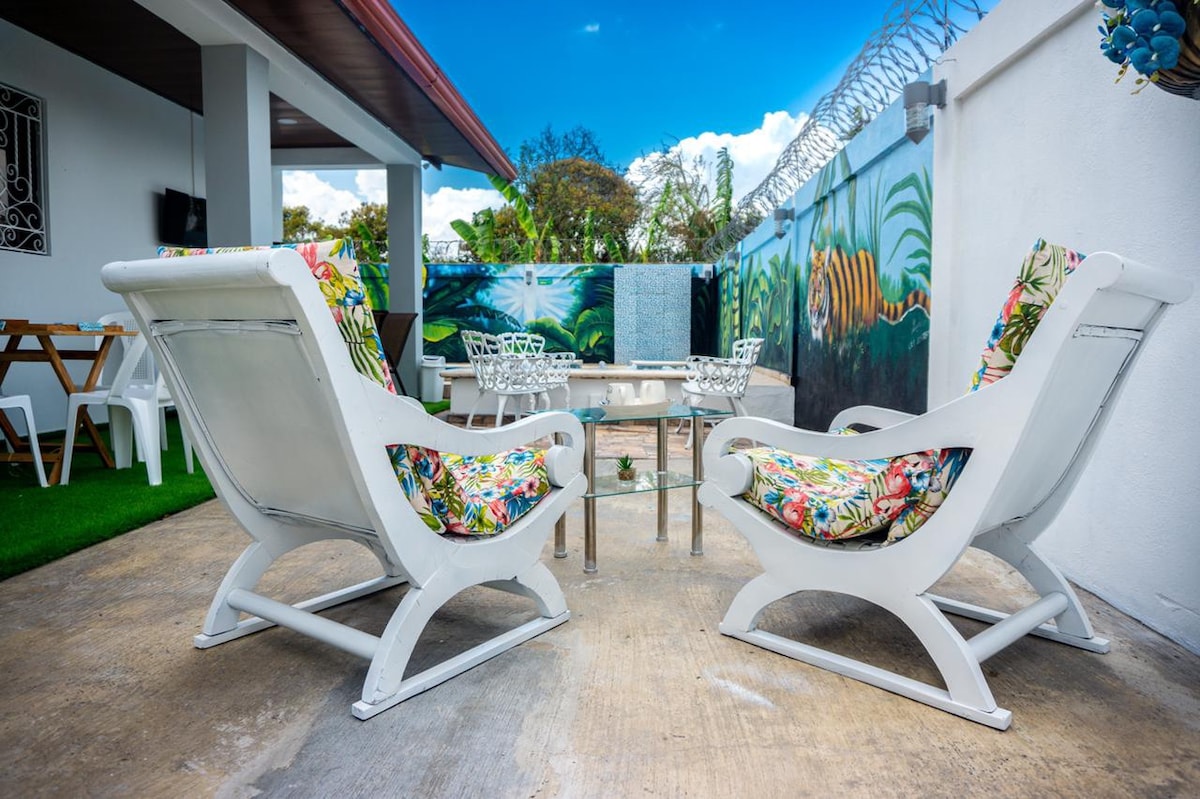 Two comfortable lounge chairs are positioned beside a small glass table in a relaxing outdoor area. Vibrant, floral cushions decorate the chairs, which face a wall mural featuring tropical foliage. A clear blue sky and greenery enhance the serene ambiance.