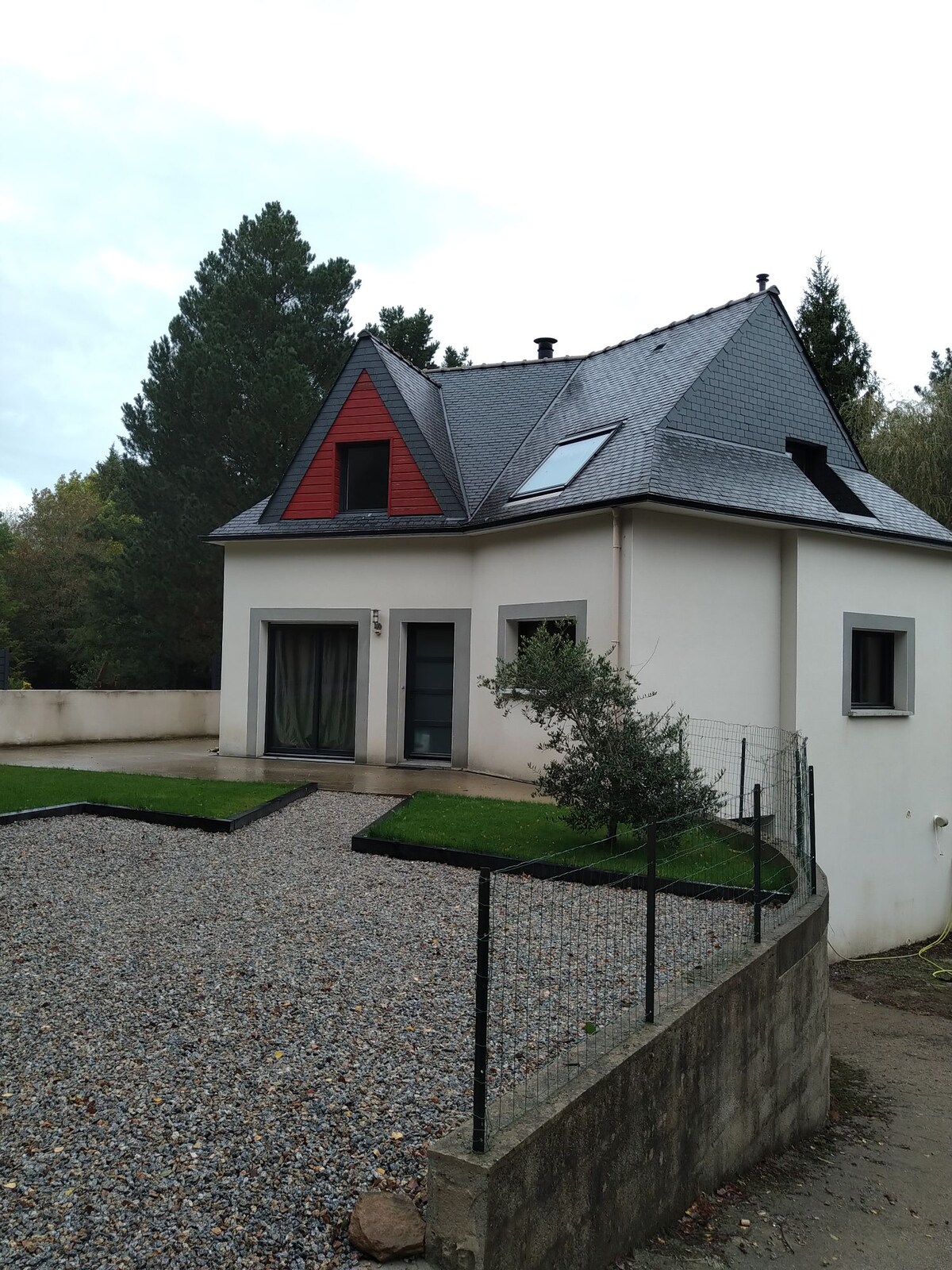 The exterior of the house features a modern design, with a grey roof and red accents. A landscaped area with gravel and patches of grass surrounds the property. Trees and greenery are visible in the background, contributing to a serene environment.