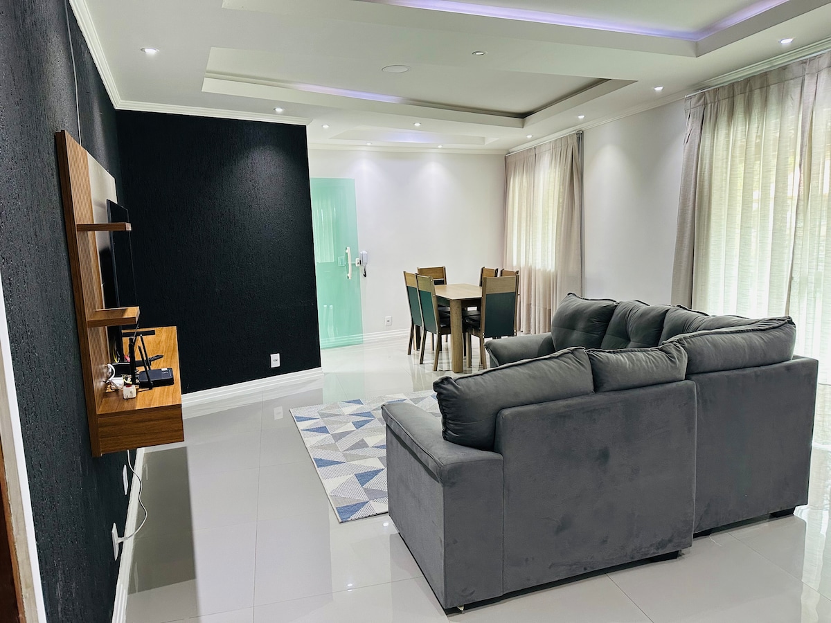 A modern living space is showcased, featuring a comfortable gray sectional sofa and a wooden TV stand. A dining area with a wooden table and chairs is visible in the background. Natural light filters through sheer curtains, illuminating the glossy tiled floor.