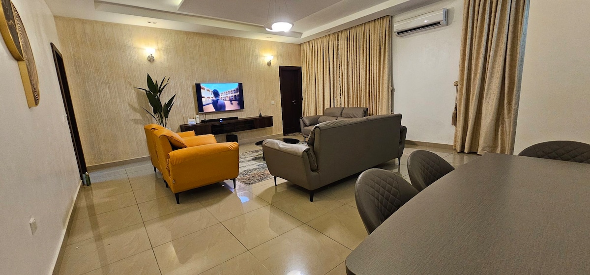 A welcoming living area features a large television mounted on a light-colored wall. A comfortable gray sofa and a bright orange chair are arranged for relaxation. Natural light enters through large windows, which are dressed with soft, elegant curtains.