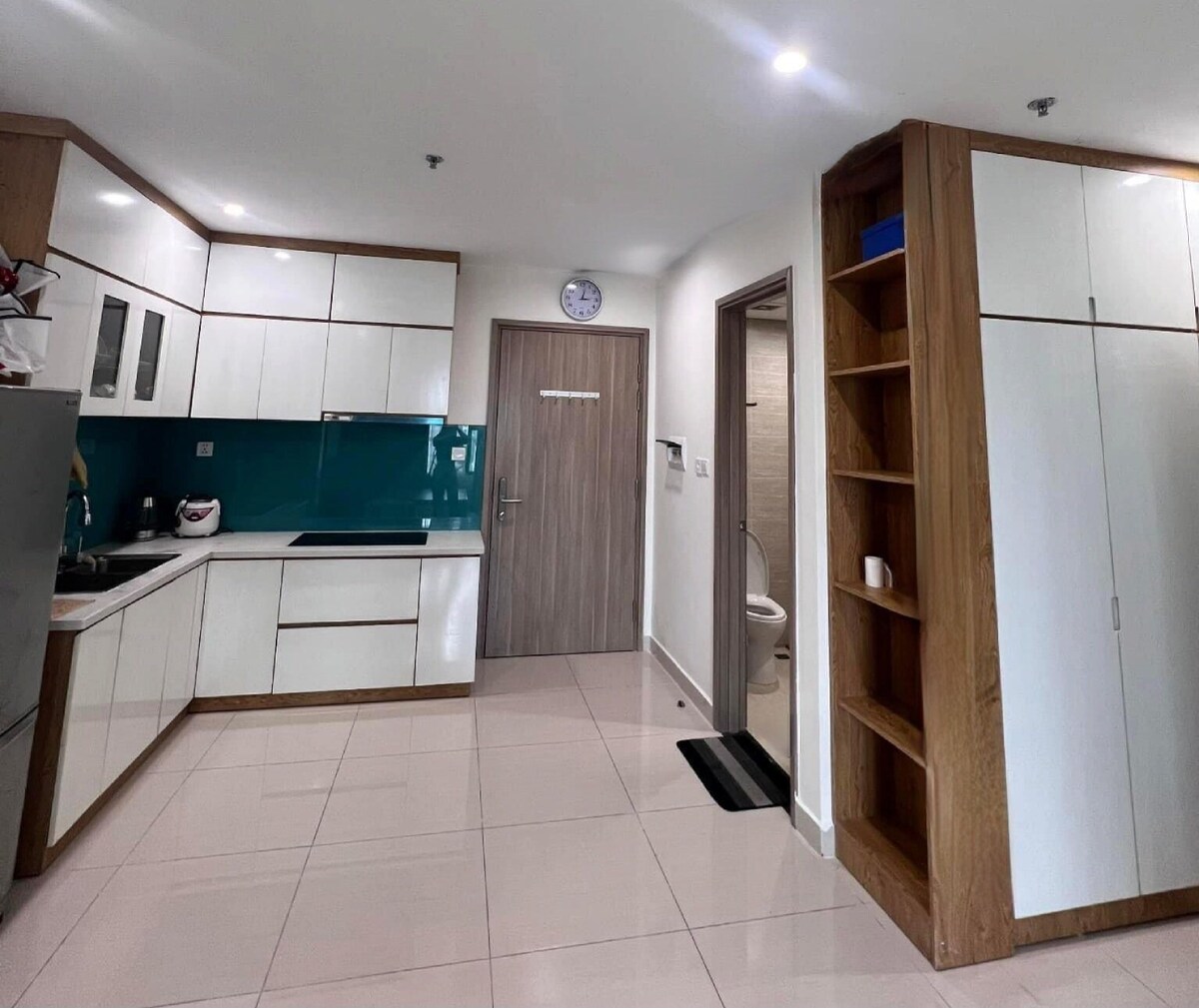 A modern kitchen is displayed, featuring sleek white cabinetry and a vibrant blue backsplash. The space is equipped with stainless steel appliances, a wooden shelf for storage, and a neutral tiled floor. A doorway leads to an adjacent bathroom, and natural light brightens the area.