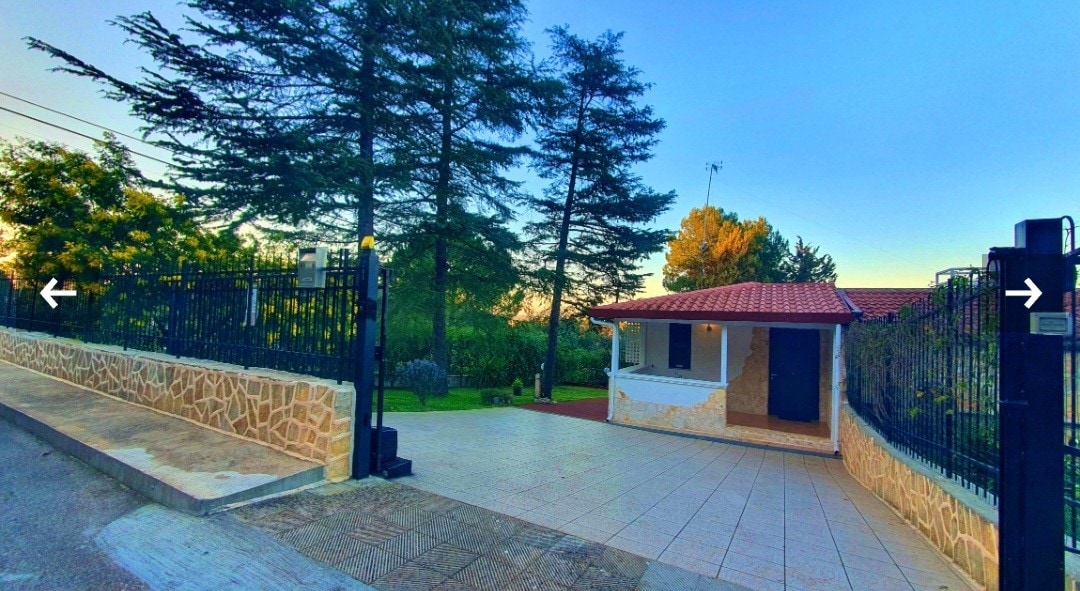 A welcoming entrance features a gated access point leading to a single-story home. The exterior displays a stone facade, complemented by a red-tiled roof. Surrounding greenery adds to the serene atmosphere, while a tiled pathway invites guests toward the front door.