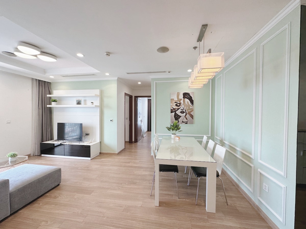 A modern living area features a light green accent wall and sleek wood flooring. A glass dining table with four chairs is positioned near a stylish artwork. A flat-screen television is mounted on a white entertainment unit, while soft natural light fills the space.
