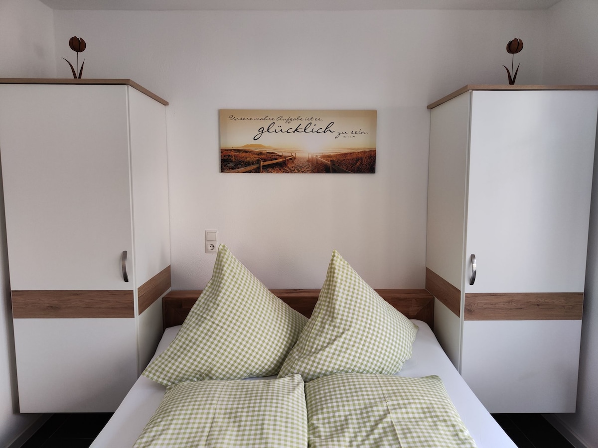 A cozy sleeping area features a spacious bed with checkered green and white pillows. Two wardrobes are positioned on either side, complemented by simple decor atop them. A serene landscape artwork hangs above the bed, adding a touch of warmth to the room.