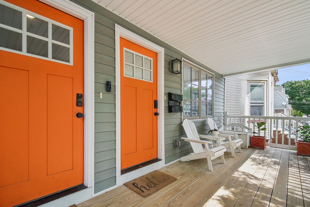 A welcoming entryway is featured with two vibrant orange doors, each equipped with a keypad entry. The porch showcases wooden flooring and two white adirondack chairs. Potted plants sit alongside the entrance, enhancing the home’s inviting atmosphere.