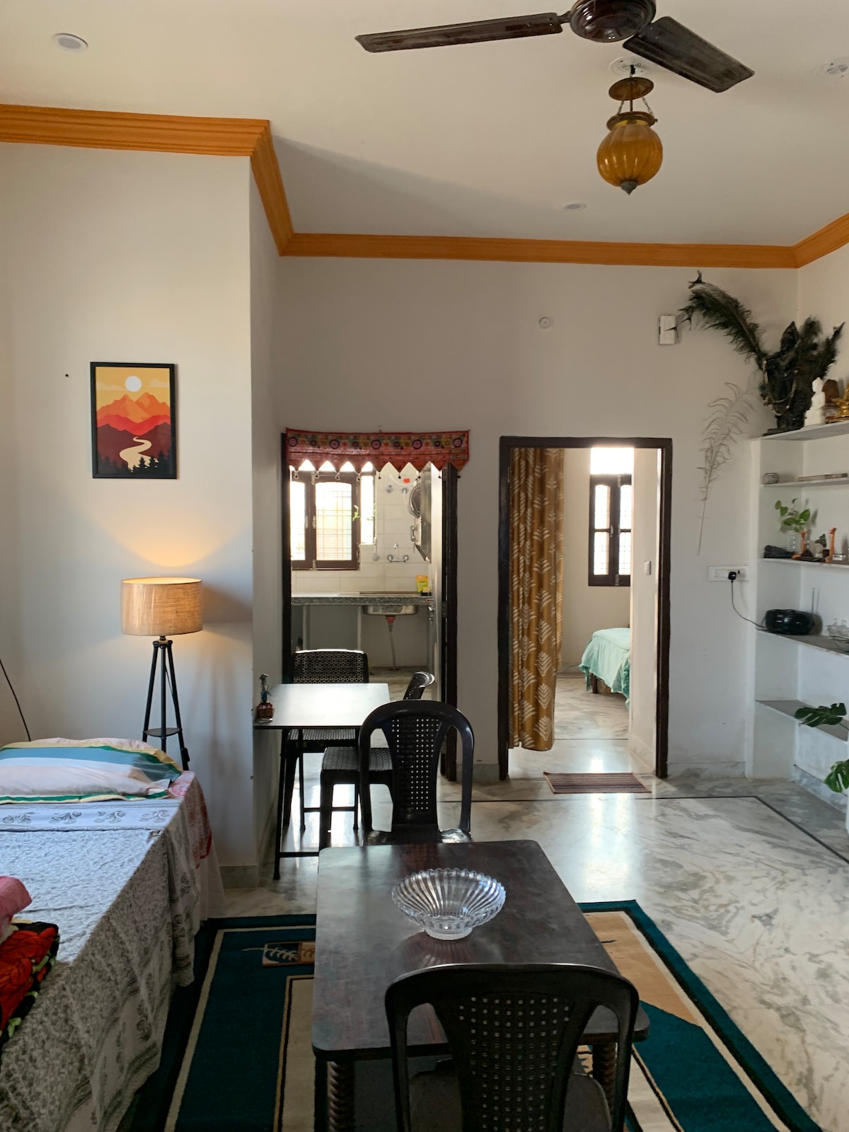 An open living and dining area features a round dining table with black chairs and a small lamp beside it. A partition door leads to a simple kitchen, while a green-tinted bedroom is visible in the background. Decorative items and plants adorn the shelves.