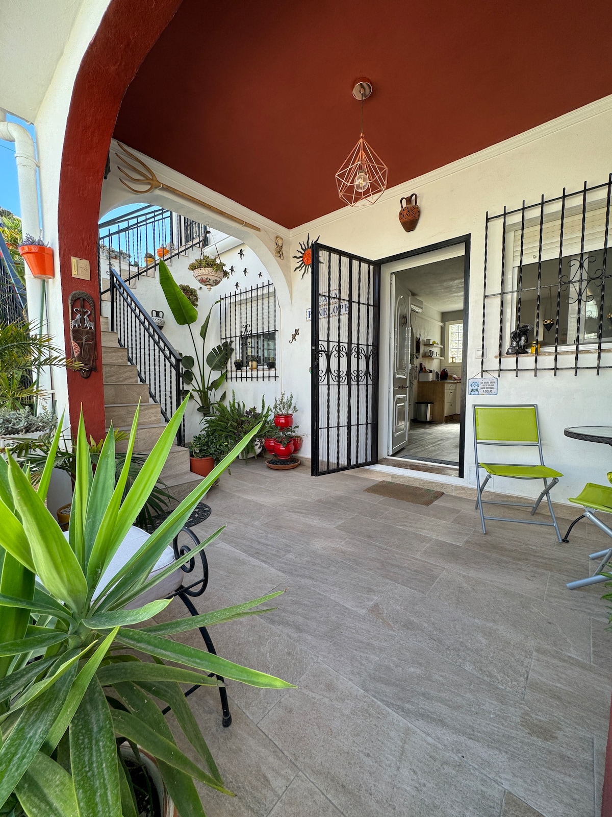 An entrance area features a decorative iron gate leading to an open kitchen. Potted plants add greenery to the space, complemented by a small dining set with two chairs and a table. A staircase is visible in the background, framed by vibrant wall colors.