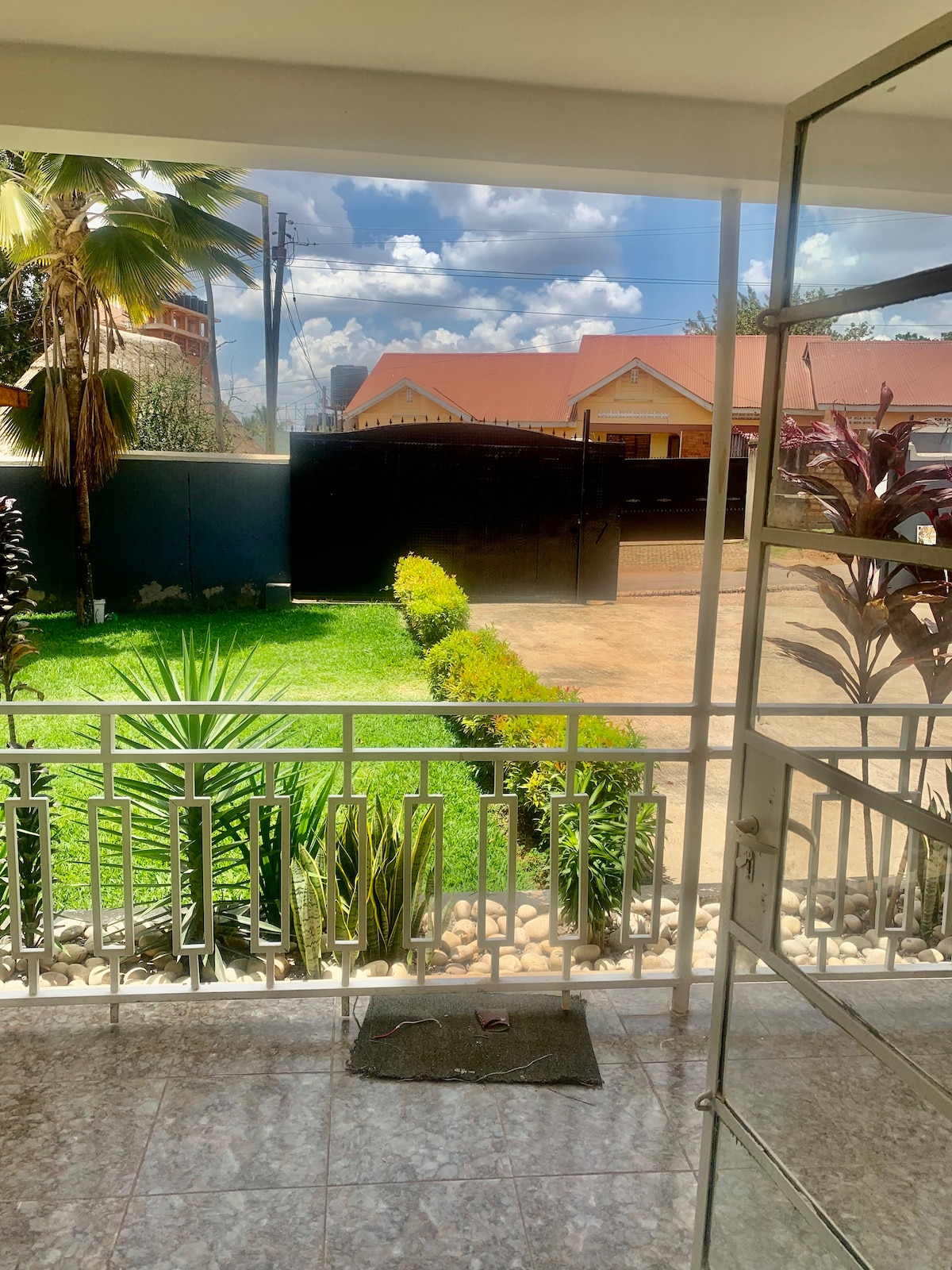 A view from the porch showcases a well-maintained garden featuring various shrubs and plants. The green lawn is framed by a stone pathway, and a gated entrance is visible in the background, providing an inviting sense of outdoor space.